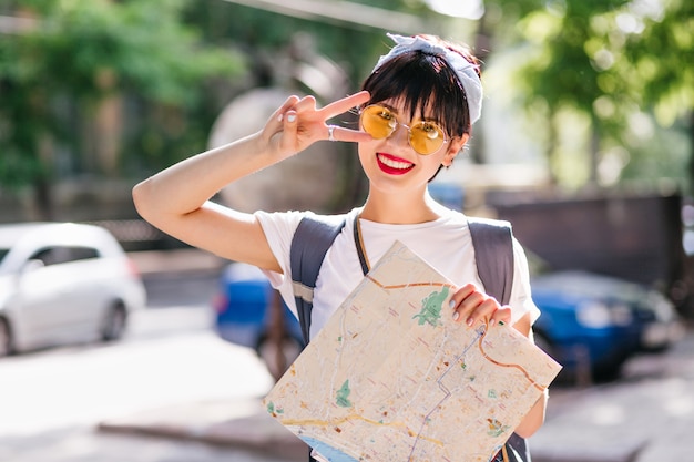 カラフルな車の前に立っているピースサインでポーズをとって魅力的な笑顔で幸せな女性旅行者