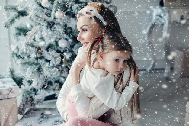 Счастливая праздничная мать и дочь обнимаются, сидя вместе за украшением красивой елки