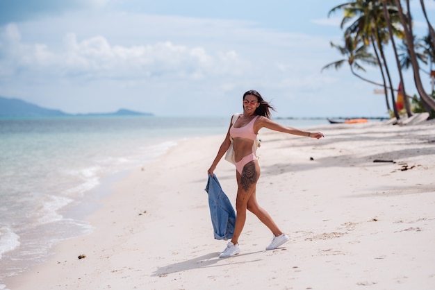 Довольно загорелая спортивная татуированная женщина счастлива подходит на пляже в джинсовой куртке