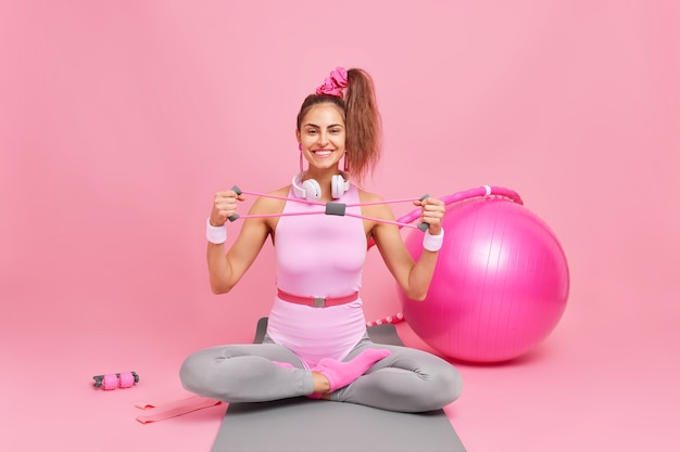 Happy fitness female model sits crossed legs on mat stretches expander trains muscles dressed in bodysuit surrounded by swiss ball resistance band hula hoop does exercises for losing weight.