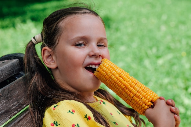 A happy fiveyearold girl nibbles corn sitting on a bench in the park on a summer day delicious boile...