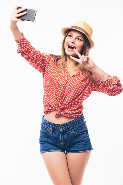 Happy flirting young woman taking pictures of herself through cell phone, over white background