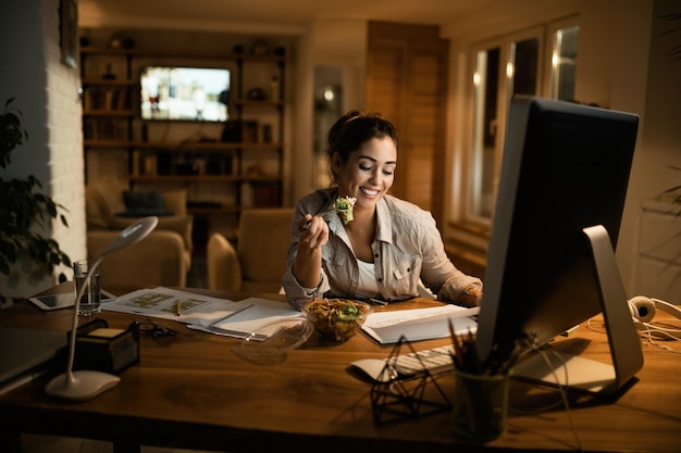 Happy freelance worker going through paperwork while eating salad in the evening at home