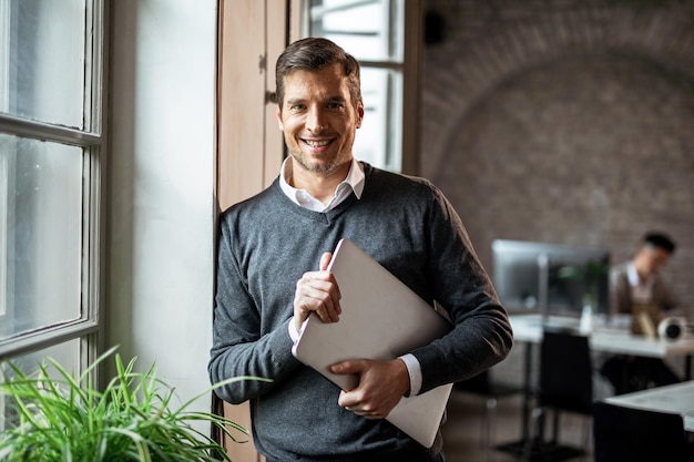 Happy freelance worker with laptop looking at camera while standing by the window in the office