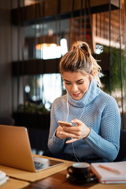 Happy freelancer using mobile phone and listening music over earphones while working on coffee break in a cafe