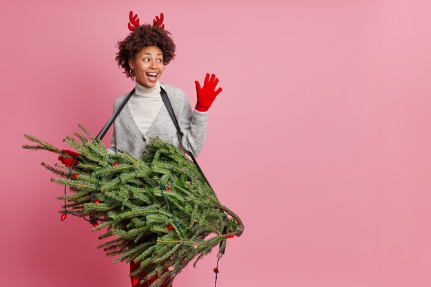 Happy friendly african american woman meets best friend on new year party foolishes around holds green christmas fir tree as if guitar pretends to be professional rock singer poses indoor blank space