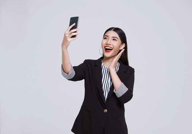 幸せでフレンドリーな顔のアジアの実業家は、スマートフォンを使用してフォーマルなスーツを着て笑顔で、白い背景のスタジオショットでビデオ通話をしています。