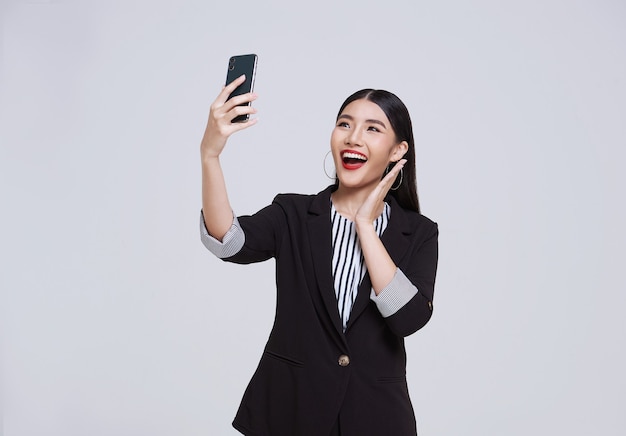 幸せでフレンドリーな顔のアジアの実業家は、スマートフォンを使用してフォーマルなスーツを着て笑顔で、白い背景のスタジオショットでビデオ通話をしています。