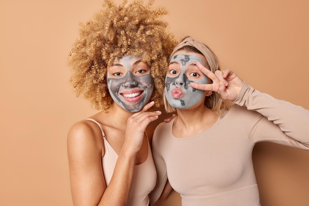 Happy friendly two women pose indoor with facial clay masks smile and make peace gesture over eye take care of skin and complexion dressed casually isolated over brown background Beauty time