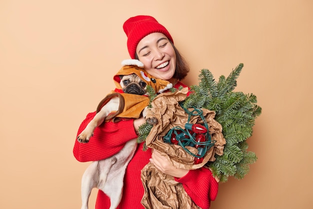 Free photo happy friendly woman embraces pug dog with love wearsred hat and jumper carries attributes for decoration feels glad has festive mood isolated over brown background. winter holidays concept.