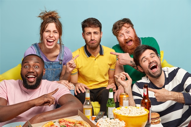 Happy friends point at unhappy bearded guy who is not eager to watch comedy, expresses dissatisfaction. Five multiethic young people have tasty snack and drink cold beer while watching stream TV