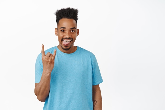 Happy and funny african american guy showing rock n roll, heavy metal horns gesture, smiling excited, enjoying music, party event, looking thrilled at camera, white background