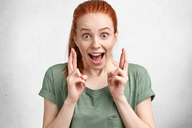 Happy funny overjoyed ginger female with hair knot, wears casual t shirt, crosses fingers with great hope, models in studio against white