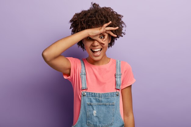 Happy funny woman looks through zero or okay gesture, holds rounded fingers near eye, smiles positively, peeps at something, feels overjoyed, wears pink t shirt and denim overalls, models indoor