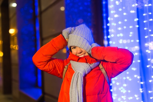 Счастливая смешная молодая женщина с зимней одеждой на фоне вечерних огней города рождество