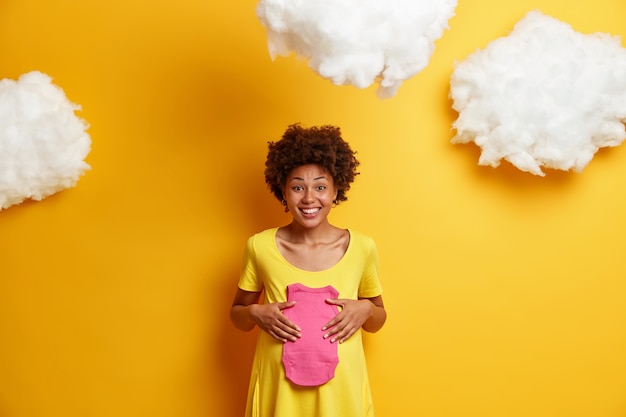 Happy future mother with pregnant belly, holds rosy singlet for unborn daughter, anticipates for baby, wears yellow dress, white fluffy clouds above. motherhood, expectation and pregnancy concept