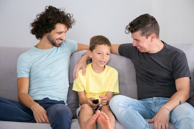 Happy gay dads and son watching an discussing TV show together at home, sitting on couch in living room, smiling and hugging boy. Family and home entertainment concept