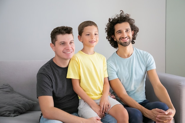 Happy gay fathers and child sitting on couch at home, smiling, and looking away. Front view. Family and parenthood concept