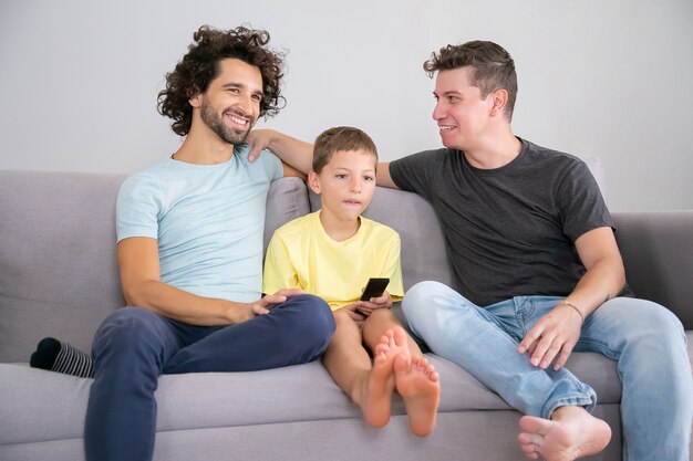 Happy gay fathers and son sitting together on couch at home, smiling, talking and looking away. Boy watching TV with remote control. Family and parenthood concept