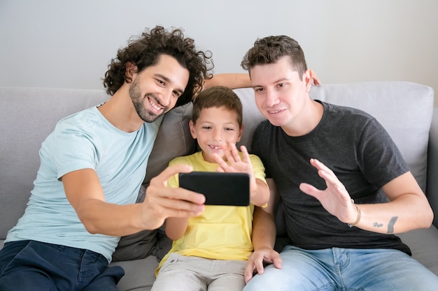 Happy gay parents and kid using cellphone for video call, sitting on couch at home, waving at frontal camera and smiling. Front view. Family and communication concept