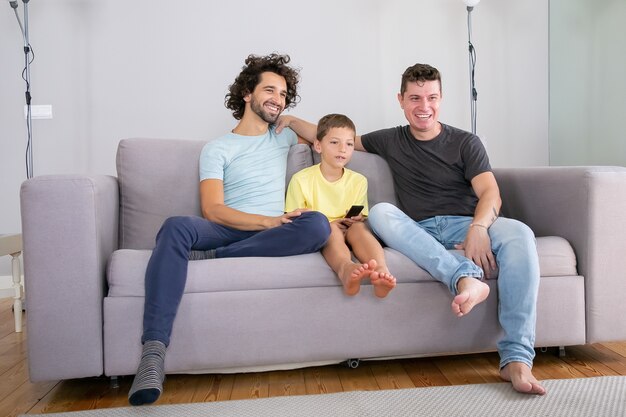 Happy gay parents and son sitting together on couch at home and watching comedy on TV, looking away, smiling and laughing. Family and parenthood concept