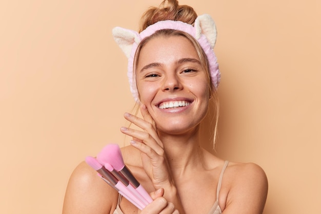 Happy gentle European woman has healthy skin touches face gently holds cosmetic brushes going to apply foundation wears headband smiles broadly isolated over brown background Beauty concept