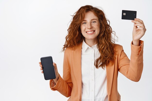 Happy ginger girl in suit showing credit card mobile phone screen demonstrating app interface standing over white background Business people concept