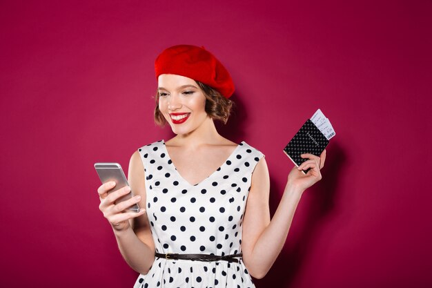 Happy ginger woman in dress holding passport with tickets and using smartphone over pink