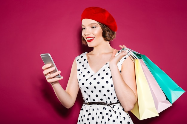 Happy ginger woman in dress with packages using smartphone over pink