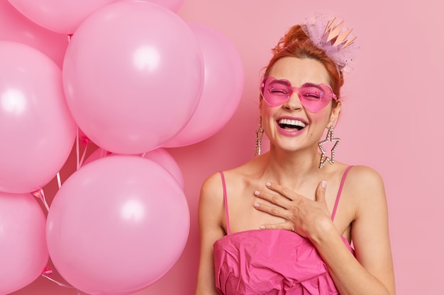 Foto gratuita la donna felice dello zenzero si sente soddisfatta sorrisi in generale tiene la mano sul petto ha umore festivo tiene palloncini gonfiati arriva alla festa di laurea