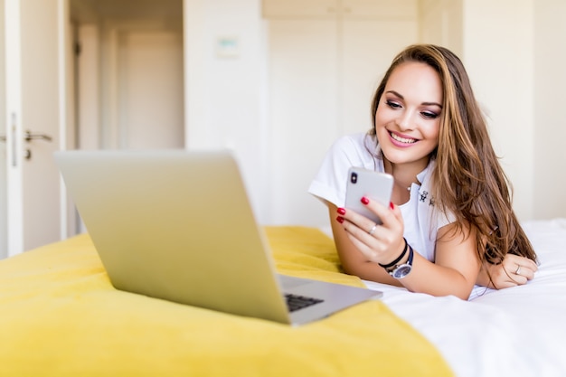 Happy girl browsing on the phone lying in bed while using laptop