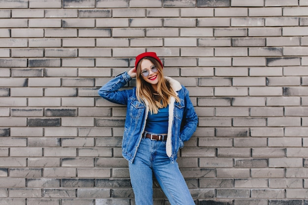 벽돌 벽 앞에 서있는 데님 복장에 행복 한 소녀. 백인 젊은 아가씨의 야외 사진은 긍정적 인 감정을 표현하는 청바지와 빨간 모자를 착용합니다.