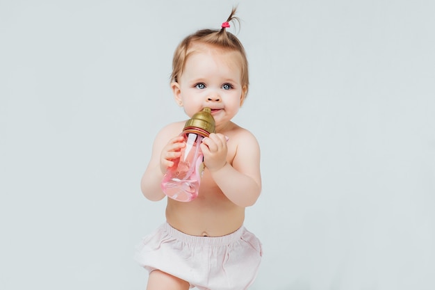 Happy girl in diaper with cute hairstyle, holds bottle of milk