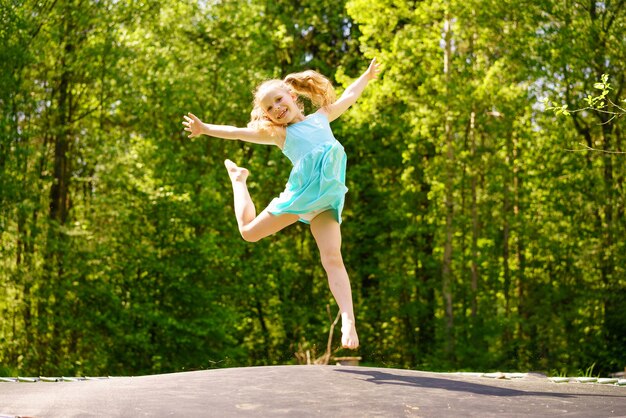 드레스를 입은 행복한 소녀가 화창한 여름날 공원에서 트램폴린에서 점프합니다.