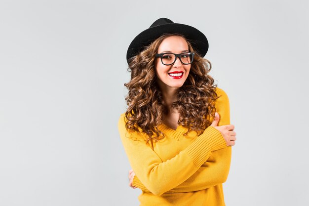 La ragazza felice in occhiali e cappello sulla parete grigia dello studio