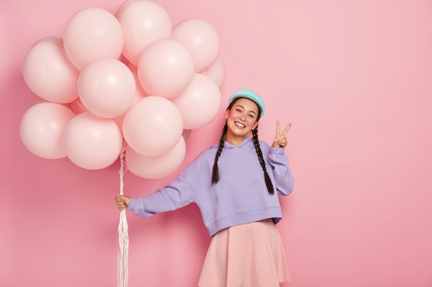 Happy girl greets friends on balloon party, has two plaits, wears purple sweater and skirt, makes peace gesture, stands against pink wall