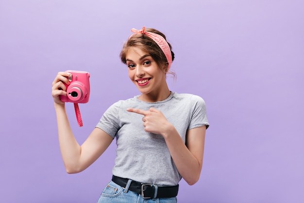 Happy girl in grey shirt pointing to pink camera. Wonderful cute woman with stylish hairstyle in grey t-shirt and denim skirt with wide belt poses.