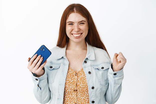 Счастливая девушка держит мобильный телефон и празднует, выглядя удовлетворенной, достигнув цели в приложении для смартфона на белом фоне