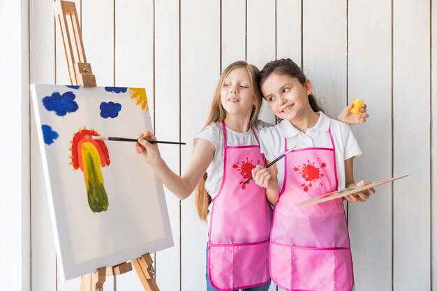 Happy girl holding palette in hand looking at her friend painting on the canvas with brush