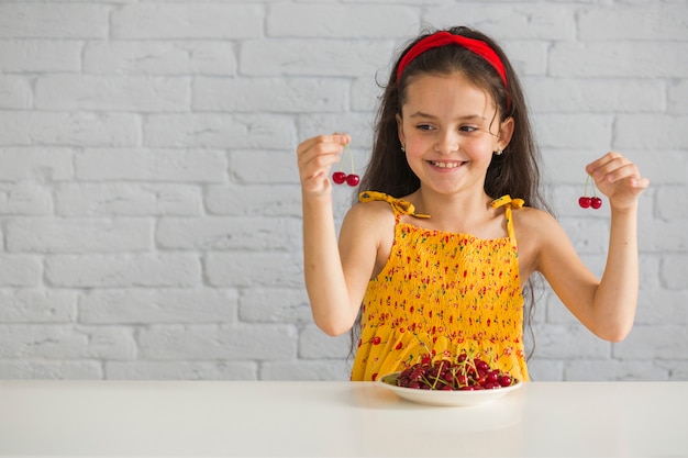 Happy girl holding red cherries on table against white brick wall