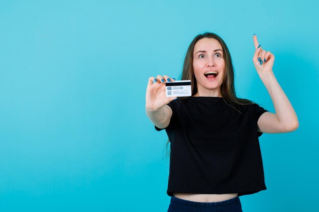 Happy girl is pointing up with forefinger and showing credit card to camera on blue background