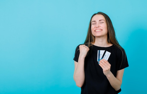 Happy girl is wishing by raising up fist and holding credit cards on blue background