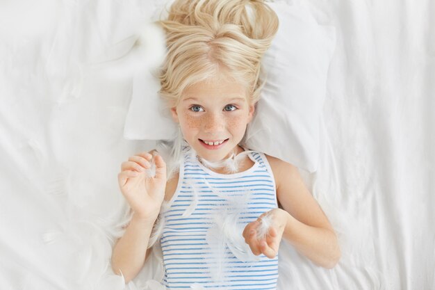 Happy girl looking with blue eyes, throwing feathers from pillow in air, having excited expression. Small naughty girl not wanting to sleep in kindergarten. Funny little kid having cheerful look