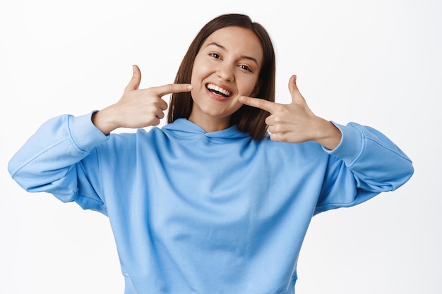 Foto gratuita ragazza felice che punta le dita al sorriso perfetto bianco, denti dritti sbiancati, in piedi in felpa con cappuccio su sfondo bianco