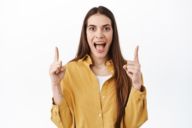 Happy girl screams and showing awesome promo deal on top, pointing fingers up and smiling broadly, demonstrating somethng cool, standing over white wall