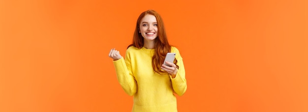 Free photo happy girl share good news with you cheerful pretty redhead female holding smartphone fist pump in t