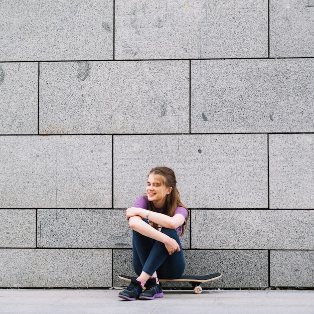 Happy girl sits on a skateboard in front of a brick wall