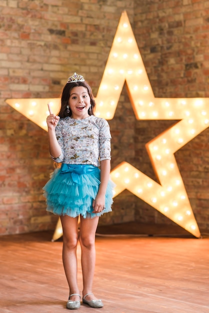 Free photo happy girl standing in front of illuminated star against brick wall pointing finger upward