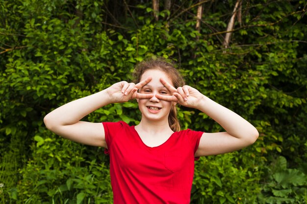 Happy girl standing in park and looking through v sign