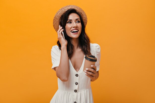 Happy girl in straw hat is talking on phone and holding glass of coffee. Beautiful lady in bright clothes holds smartphone and tea on isolated background.